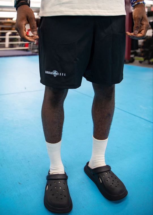 Person wearing black shorts with a logo, white socks, and black Crocs in a boxing ring.